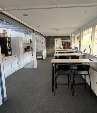 Leefruimte met hoge tafels, stoelen en een open keuken aan de rechterkant.